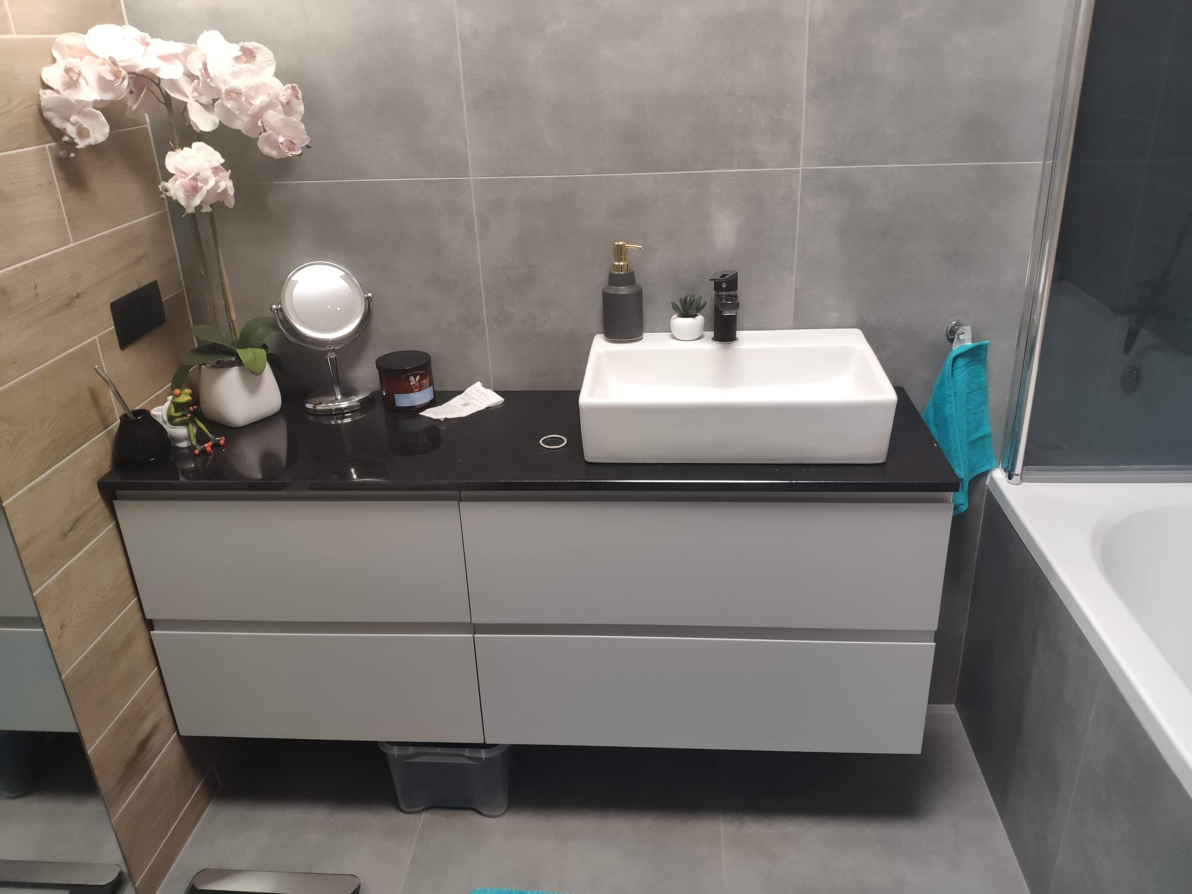 Nowoczesna łazienka z szarymi płytkami, białą umywalką nablatową na szafce z szufladami, czarnym blatem i dekoracjami: sztuczne kwiaty, lusterko, dozownik mydła i mała roślina w doniczce.