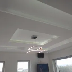 Biały sufit podwieszany z geometrycznym wzorem i oświetleniem LED, nowoczesna lampa wisząca z kwadratowymi elementami, widok z dołu na wykończone wnętrze salonu z oknami.