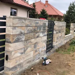 Fragment ogrodzenia z bloków betonowych o nieregularnym wzorze i stalowej bramki wejściowej, widoczny numer domu '5W' oraz narzędzia budowlane na ziemi.