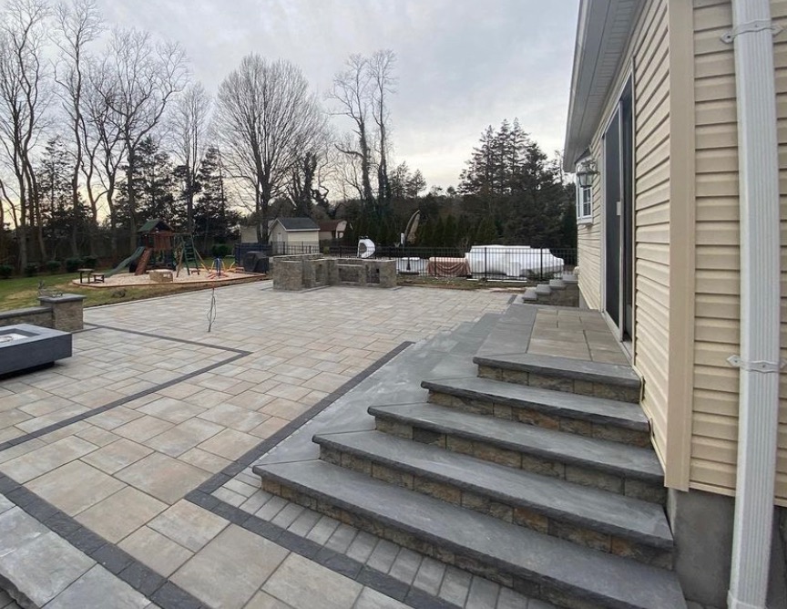Wyłożony kostką brukową taras z kamiennymi schodami prowadzącymi do domu z sidingiem, w tle plac zabaw i zabudowa grilla
