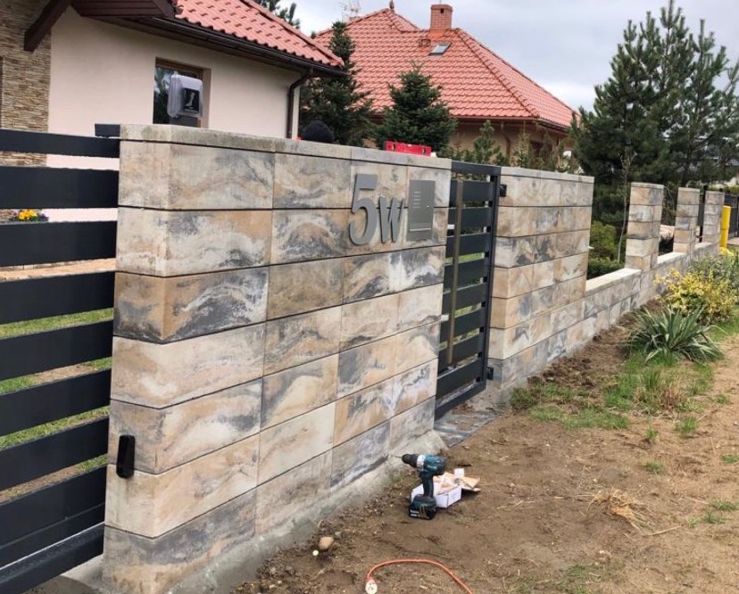 Fragment ogrodzenia z bloków betonowych o nieregularnym wzorze i stalowej bramki wejściowej, widoczny numer domu '5W' oraz narzędzia budowlane na ziemi.