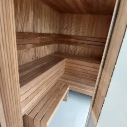 Wnętrze nowej, wykończonej sauny z drewnianymi ławkami i boazerią, widok z zewnątrz przez częściowo otwarte szklane drzwi.