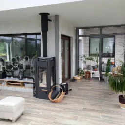 Nowoczesny, czarny piec wolnostojący z rurą odprowadzającą dym, ustawiony w przestronnym, jasnym pomieszczeniu z dużymi oknami i ceramiczną podłogą imitującą drewno, kosz wiklinowy z czarnym...