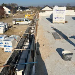 Wylewanie fundamentów pod budowę domu jednorodzinnego; widoczne szalunki zbrojeniowe, bloczki Solbet i Prefabet, oraz zaparkowany bus firmy budowlanej na tle budynków mieszkalnych.