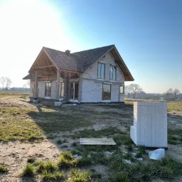 Niedokończony dom murowany z szarym dachem i drewnianą konstrukcją dachu, widoczne okna, na pierwszym planie paleta i bloczki budowlane.