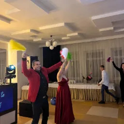 Animator w czerwonej marynarce i kobieta w czerwonej sukni z kolorowymi piórami na sali balowej, w tle osoby i oświetlenie eventowe.