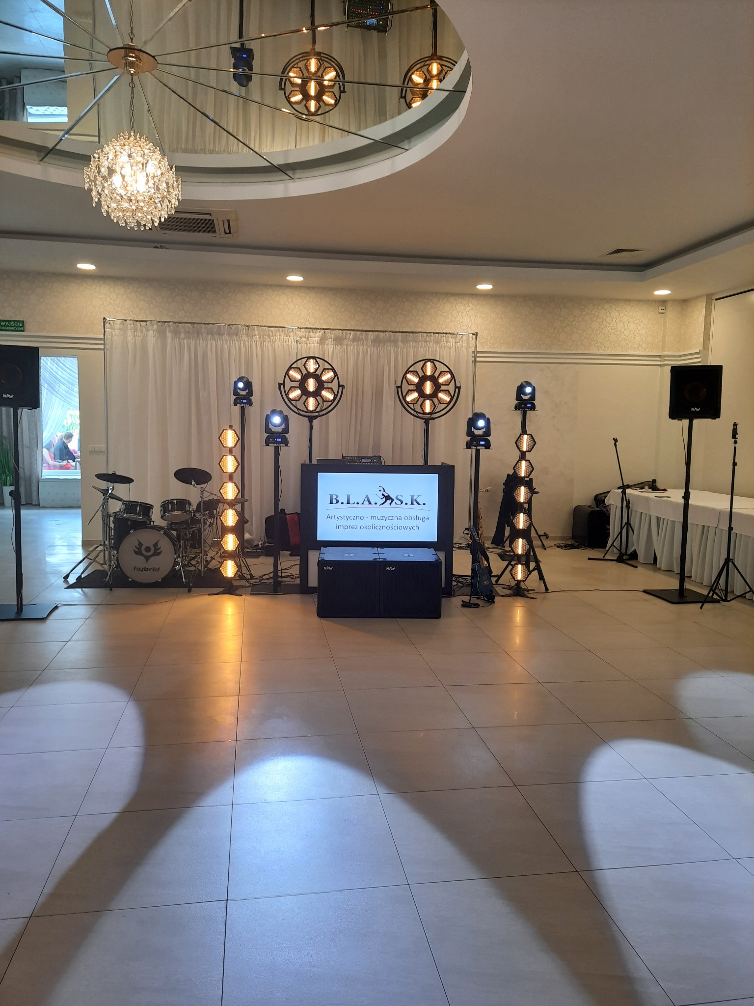 Sprzęt nagłośnieniowy i oświetleniowy zespołu muzycznego B.L.A.S.K. na sali balowej. Widoczny zestaw perkusyjny, kolumny, reflektory i telebim z nazwą zespołu.