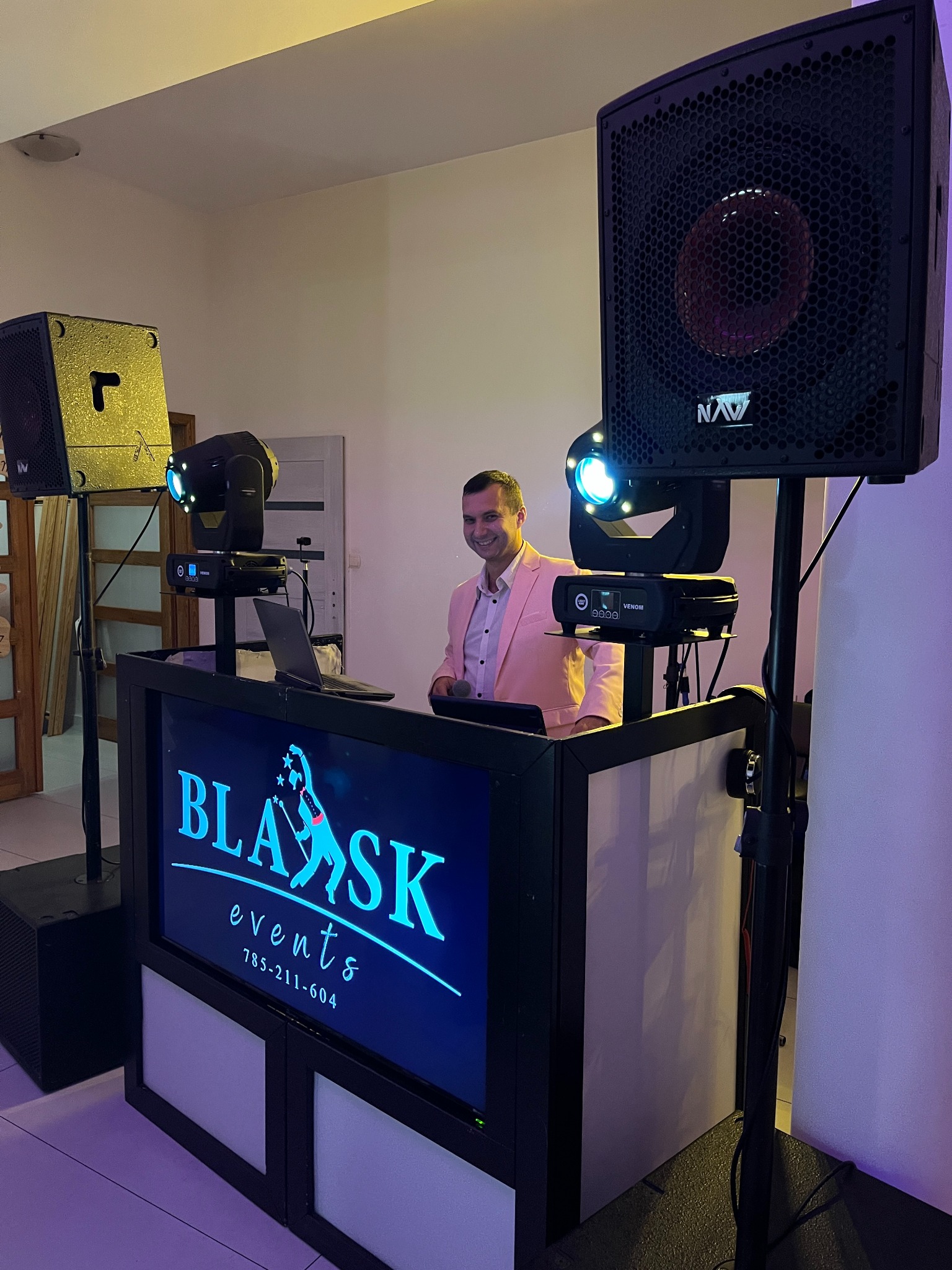 Uśmiechnięty DJ w różowej marynarce za stanowiskiem z logo 'Blask Events', oświetlony reflektorami i głośnikami, gotowy do pracy na imprezie.