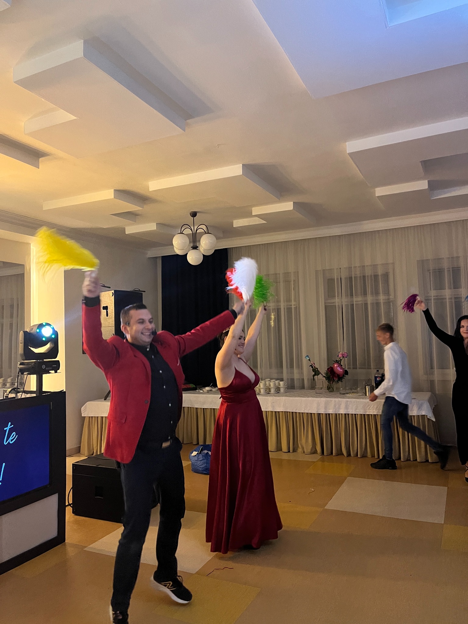 Animator w czerwonej marynarce i kobieta w czerwonej sukni z kolorowymi piórami na sali balowej, w tle osoby i oświetlenie eventowe.