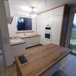 Nowoczesna kuchnia z białymi szafkami, drewnianymi blatami i wyspą kuchenną z gniazdem elektrycznym w blacie, oświetlona lampą sufitową.