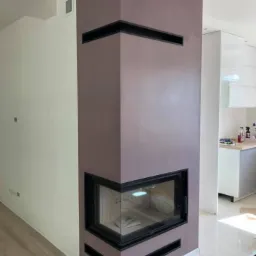 Nowoczesny kominek narożny w zabudowie w kolorze liliowym z czarnymi akcentami, widoczny fragment parkietu i kuchni w tle.