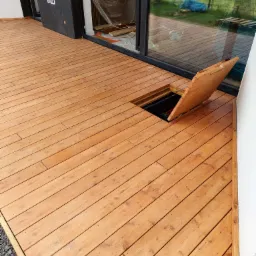 Nowy taras z desek z drewna z otwieraną klapą do schowka, widoczne kamienie obok tarasu i okno przesuwne z folią ochronną.