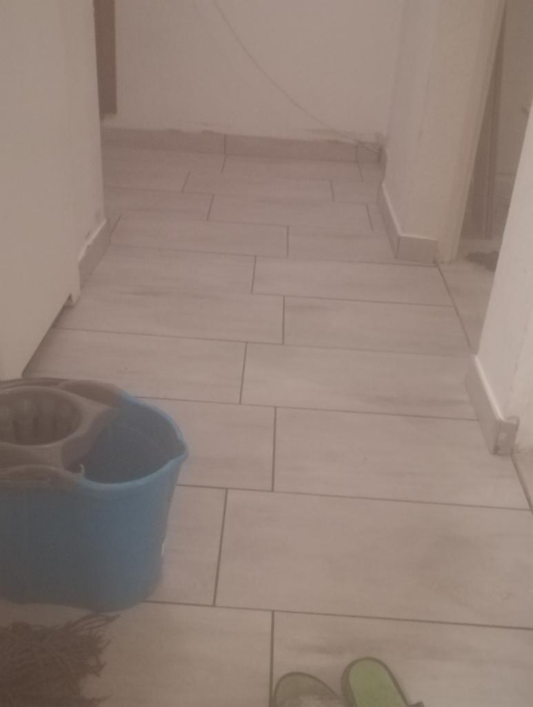 Świeżo położone, jasne płytki podłogowe w korytarzu, widoczne wiadro z mopem i fragment klapek w rogu kadru.