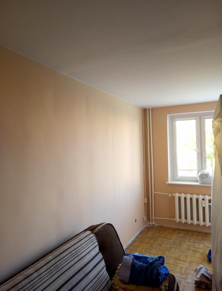 Świeżo pomalowane ściany w odcieniach beżu w pokoju z parkietem, widoczne rury grzewcze i okno z białymi ramami, częściowo zasłonięty narożnik sofy z wzorzystą narzutą.