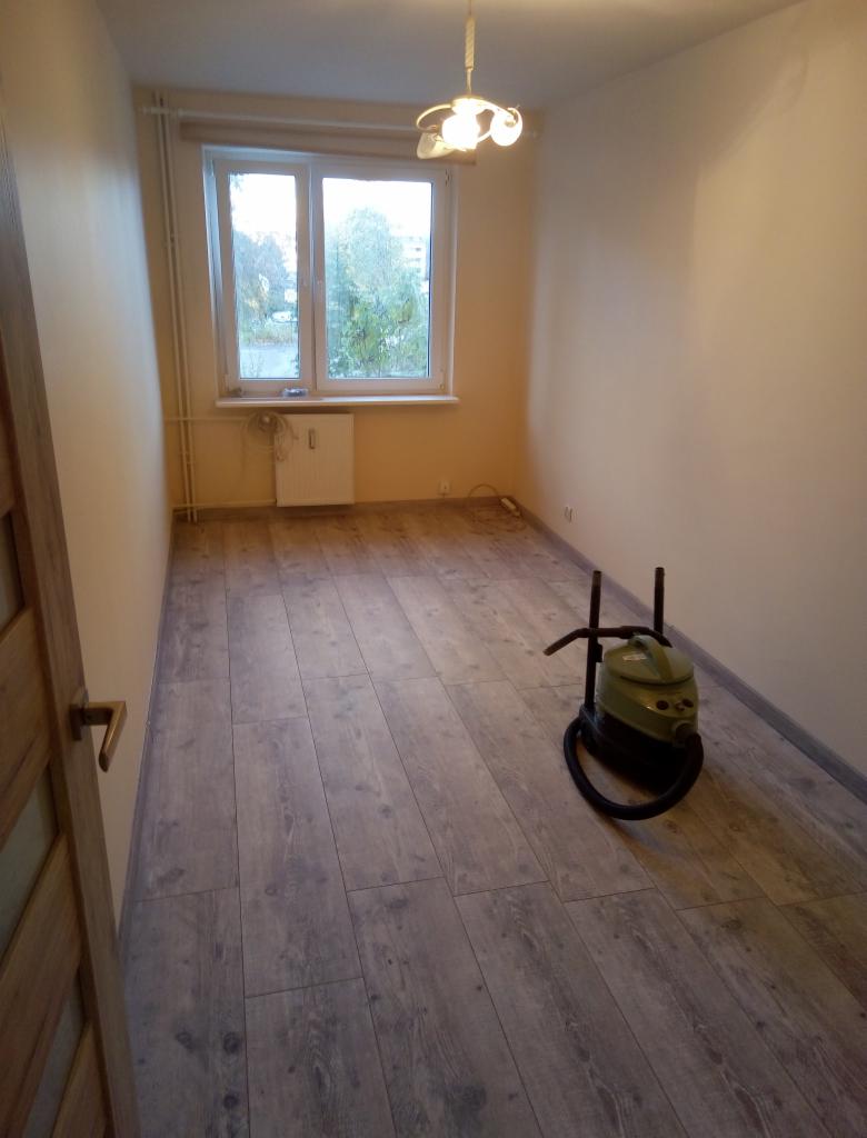 Pokój po remoncie z nową podłogą z paneli imitujących drewno, widoczny odkurzacz i okno z widokiem na zieleń.