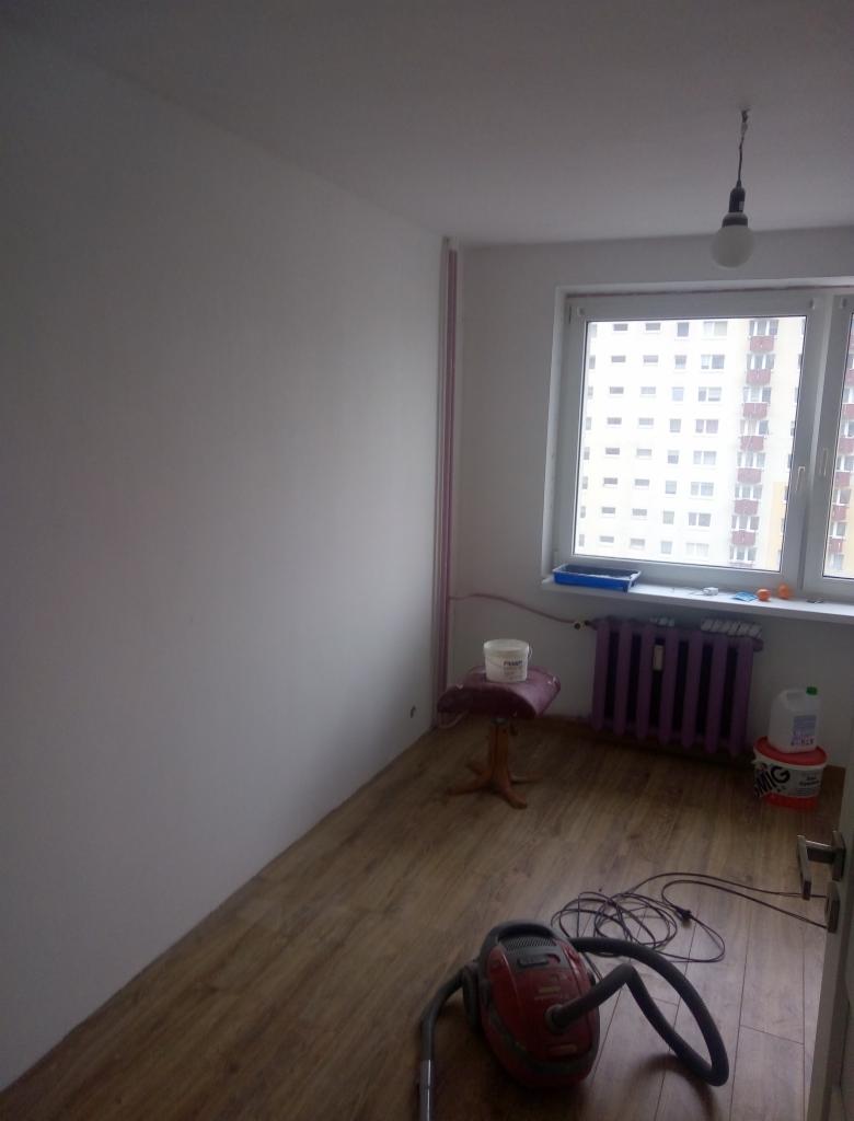 Pomieszczenie po malowaniu ścian na biało, widoczny odkurzacz, grzejnik pomalowany na fioletowo, wiadro farby i taboret.