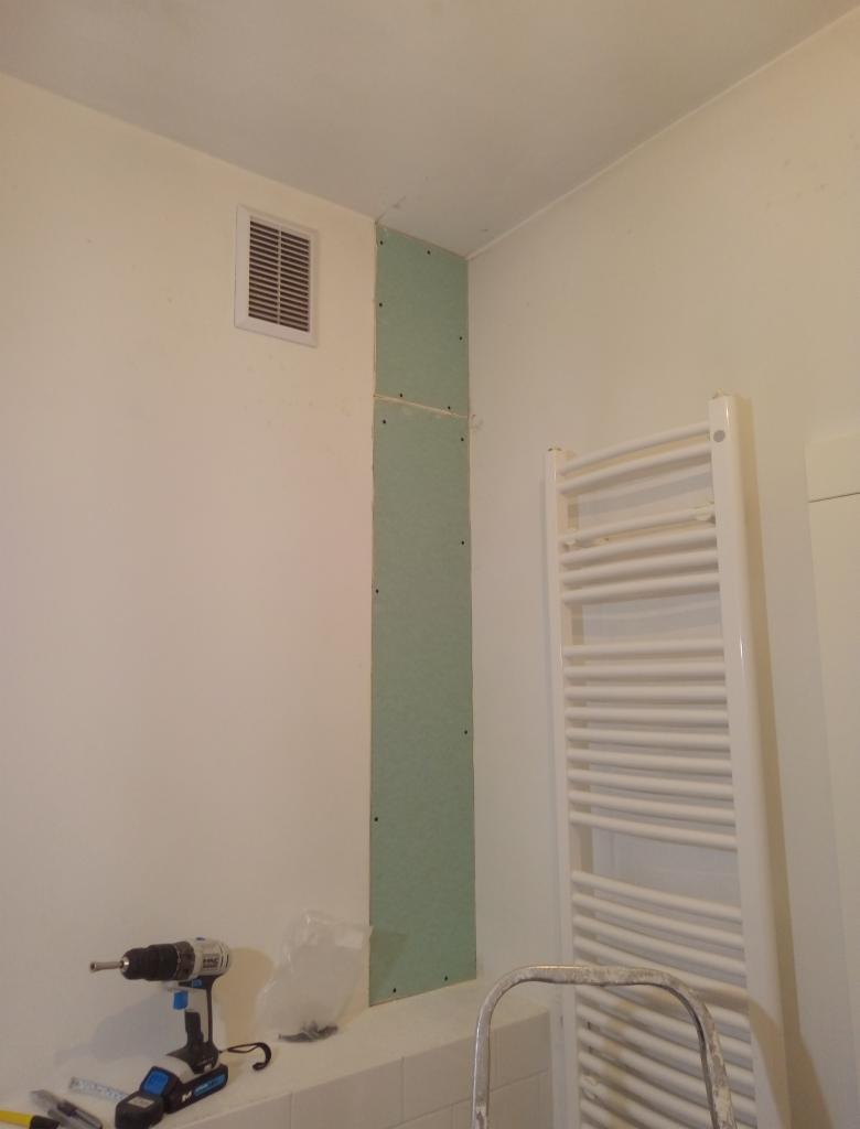Montaż zielonej płyty gipsowo-kartonowej na ścianie w łazience, widoczny grzejnik drabinkowy, wiertarka akumulatorowa i wentylator w ścianie, etap prac wykończeniowych.