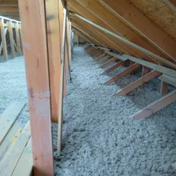 Poddasze w Kolbudach po ociepleniu wełną mineralną, widoczne drewniane krokwie i legary, oświetlenie w tle.