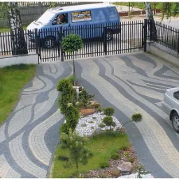 Wzorzysty podjazd z kostki brukowej w odcieniach szarości, z elementami dekoracyjnymi w postaci roślinności i białych kamieni, widok z góry na posesję z samochodem i furgonetką za ogrodzeniem.