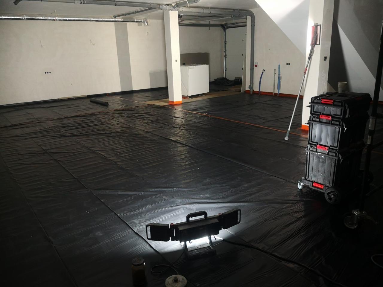 Przygotowanie pomieszczenia do prac wykończeniowych: rozłożona czarna folia ochronna na podłodze, widoczne narzędzia i oświetlenie robocze, ściany częściowo przygotowane.