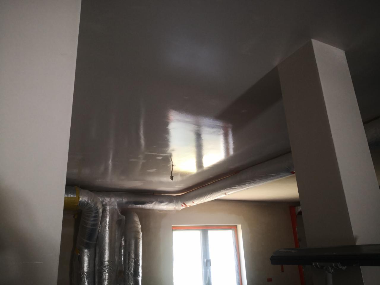 Świeżo położona, błyszcząca gładź na suficie w trakcie remontu, widoczne zabezpieczone rury wentylacyjne i kolumna konstrukcyjna.