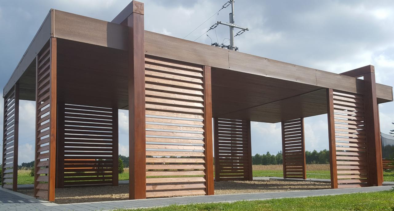 Nowoczesna, otwarta konstrukcja z ciemnego drewna z poziomymi listwami, widok z zewnątrz, na tle zieleni i pochmurnego nieba, z elementami infrastruktury energetycznej w tle.