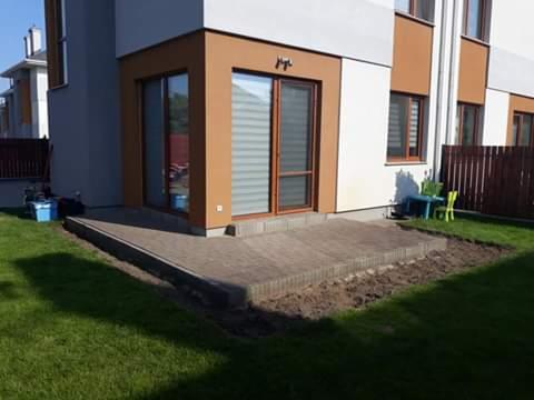 Niski taras z kostki brukowej przy nowoczesnym domu z brązowymi ramami okiennymi, widoczne przygotowane podłoże pod dalszą część tarasu, narzędzia i akcesoria budowlane po lewej stronie.