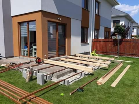 Budowa tarasu przy domu jednorodzinnym: widoczne betonowe bloczki fundamentowe, legary i deski tarasowe, narzędzia na trawie, dom z brązowymi ramami okiennymi w tle.