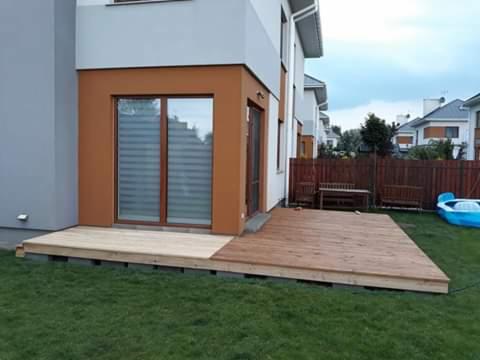 Fragment drewnianego podestu przy domu, część surowa, część zaimpregnowana, widoczny fragment trawnika i ogrodzenia.