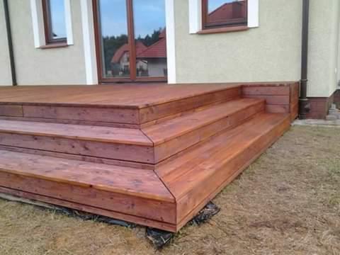 Drewniany taras z trzema stopniami prowadzący do drzwi balkonowych, widok z niskiej perspektywy na tle elewacji domu.