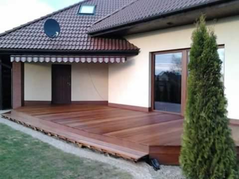 Nowoczesny taras z drewna egzotycznego przylegający do domu z brązowymi oknami, widoczna markiza nad wejściem i iglak obok tarasu.