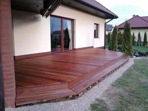 Drewniany, dwupoziomowy podest przy domu z brązowymi drzwiami przesuwnymi, otoczony trawnikiem i niskimi tujami.