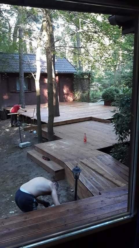 Budowa drewnianego tarasu wokół drzew w ogrodzie, widok z okna, pracownicy w trakcie pracy, butelka napoju na tarasie.