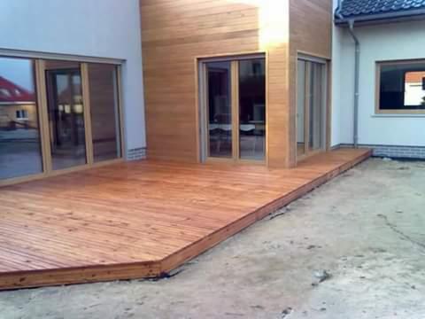 Drewniany podest przy domu z jasnymi, przesuwnymi drzwiami tarasowymi i elewacją z jasnego drewna, widoczne ślady budowy.