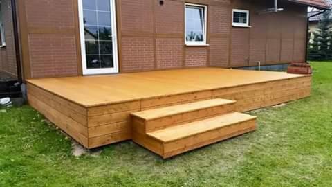 Drewniany taras z trzema schodkami prowadzącymi do drzwi, umiejscowiony przed domem z brązową elewacją.