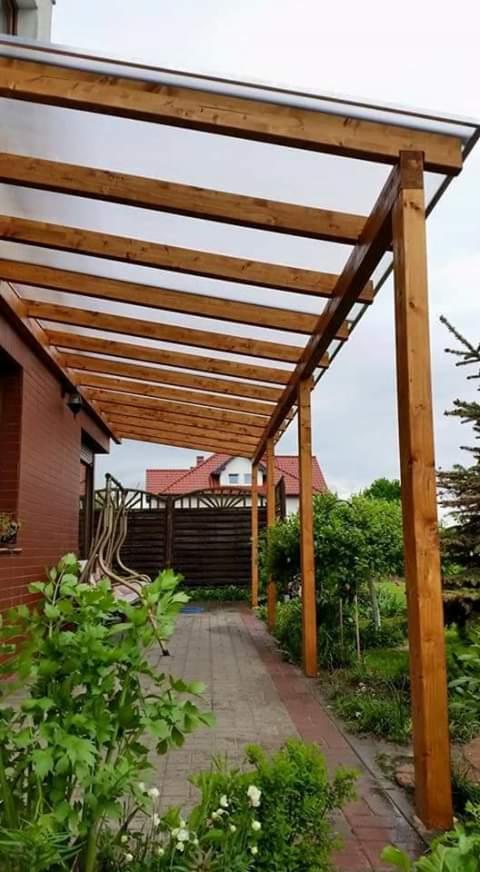 Drewniana pergola przylegająca do ceglanego domu, z przezroczystym zadaszeniem i widocznym ogrodem z kostką brukową.