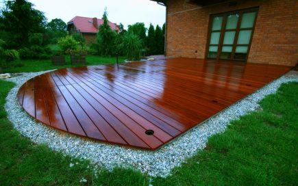 Półokrągły taras z drewna egzotycznego, obramowany białym grysem, przylegający do ceglanego budynku z dużym oknem.