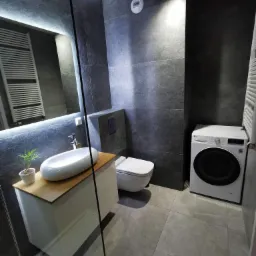 Nowoczesna łazienka z szarymi płytkami imitującymi kamień, podświetlanym lustrem, umywalką nablatową na drewnianym blacie, toaletą podwieszaną i pralką LG.