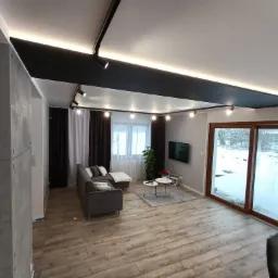 Nowoczesny salon z szarą sofą narożną, drewnopodobną podłogą, czarnym sufitem podwieszanym z oświetleniem punktowym i dużym oknem z widokiem na zimowy krajobraz.