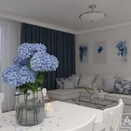 Wnętrze salonu z narożną sofą w odcieniach bieli i szarości, akcentowane niebieskimi poduszkami i obrazami. Na pierwszym planie stół z białymi krzesłami i bukietem błękitnych hortensji w szklanym...