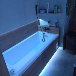 Nowoczesna łazienka w Starachowicach z podświetlaną wanną, drewnopodobnymi panelami i szarą glazurą podłogową, widoczne półki z kosmetykami i armatura.