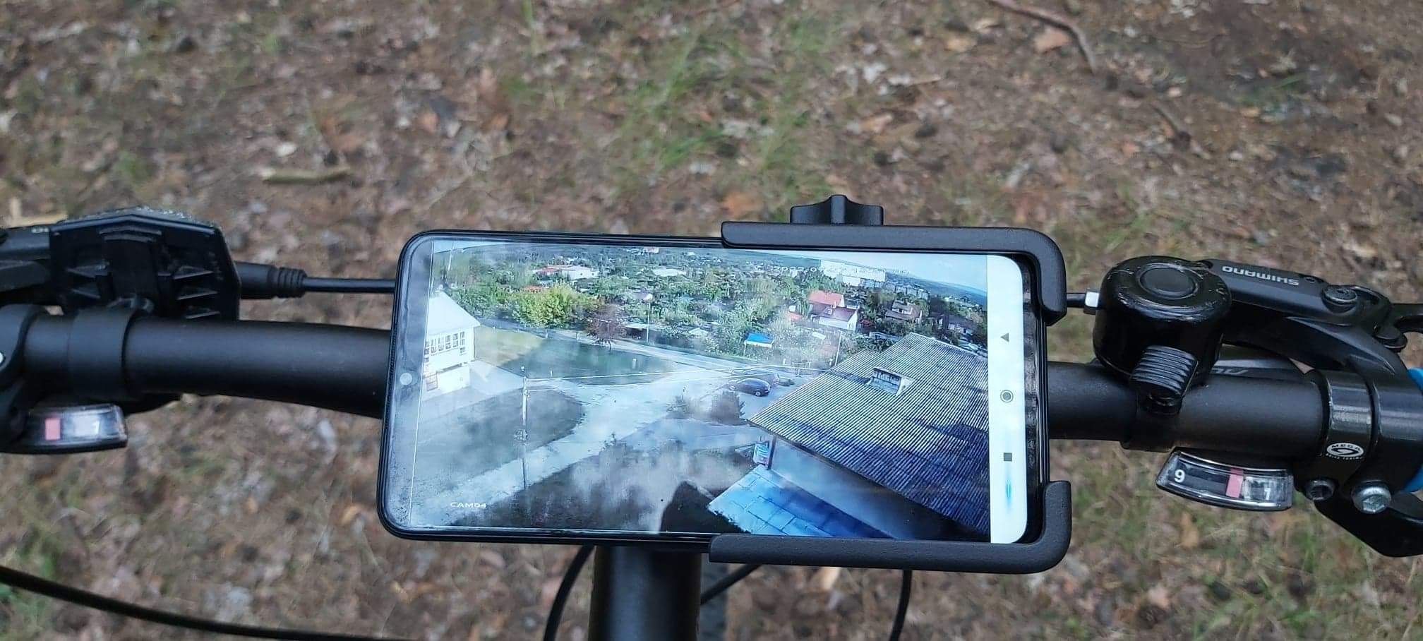 Smartfon zamocowany na kierownicy roweru wyświetlający obraz z kamery monitoringu domu, widoczny fragment dachu i otoczenie.