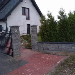 Wjazd na posesję z bramą z kutego żelaza, murowane słupki z szarej kostki, fragment murku z kostki w tle domu jednorodzinnego i zielonych tuj.