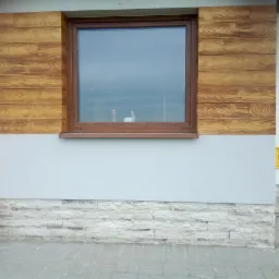 Fragment elewacji budynku z oknem, wykończony panelami imitującymi drewno na górze i kamieniem dekoracyjnym na dole, oddzielonymi gładkim, jasnym tynkiem.