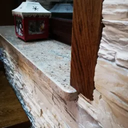 Jasny parapet z nakrapianego kamienia z dekoracyjną, świąteczną latarenką, zamontowany w oknie z drewnianą ramą i okładziną ściany imitującą kamień.