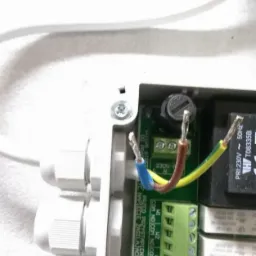 Widok z bliska panelu elektrycznego w białej obudowie z podłączonymi, odizolowanymi przewodami (niebieski, brązowy, żółto-zielony) do zielonej listwy zaciskowej. Widoczne także elementy...