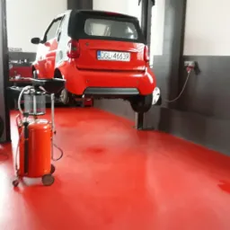 Czerwony Smart na podnośniku samochodowym w warsztacie, widoczne narzędzia i wózek serwisowy, czerwona posadzka, oświetlenie górne.