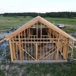 Wznoszenie szkieletu drewnianego domu z widoczną konstrukcją ścian, belek stropowych i dachu, na tle zielonego krajobrazu z lasem i polami, z widocznym rusztowaniem, paletami materiałów budowlanych...