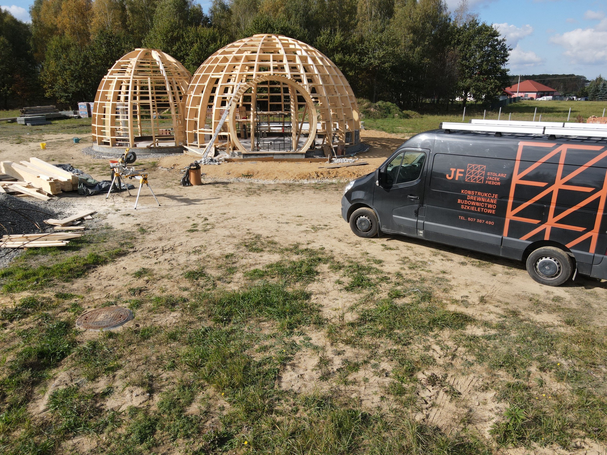 Dwa niedokończone geodezyjne domy kopułowe ze szkieletem drewnianym na działce, z widocznym samochodem dostawczym firmy budowlanej z logo i numerem telefonu.
