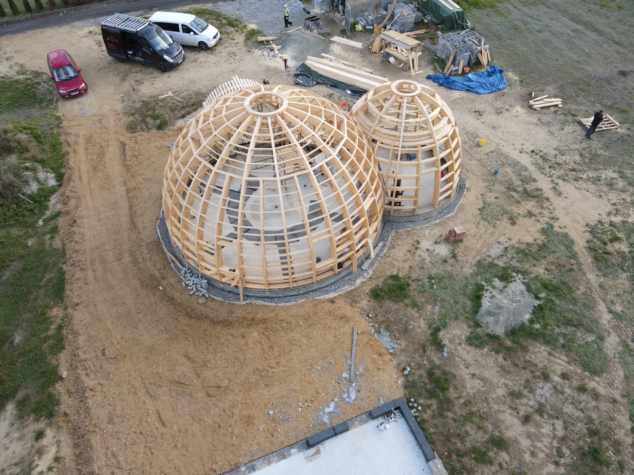 Dwa drewniane szkielety kopuł w trakcie budowy, widok z góry, na placu budowy widoczne samochody i materiały budowlane, lekko pochmurne niebo.